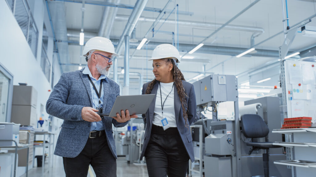 Portrait of a Two Diverse Heavy Industry Manufacturing Engineers in Hard Hats Walking with Laptop Computer and Talking in a Factory. Footage of Two Manufacturing Employees at Work.