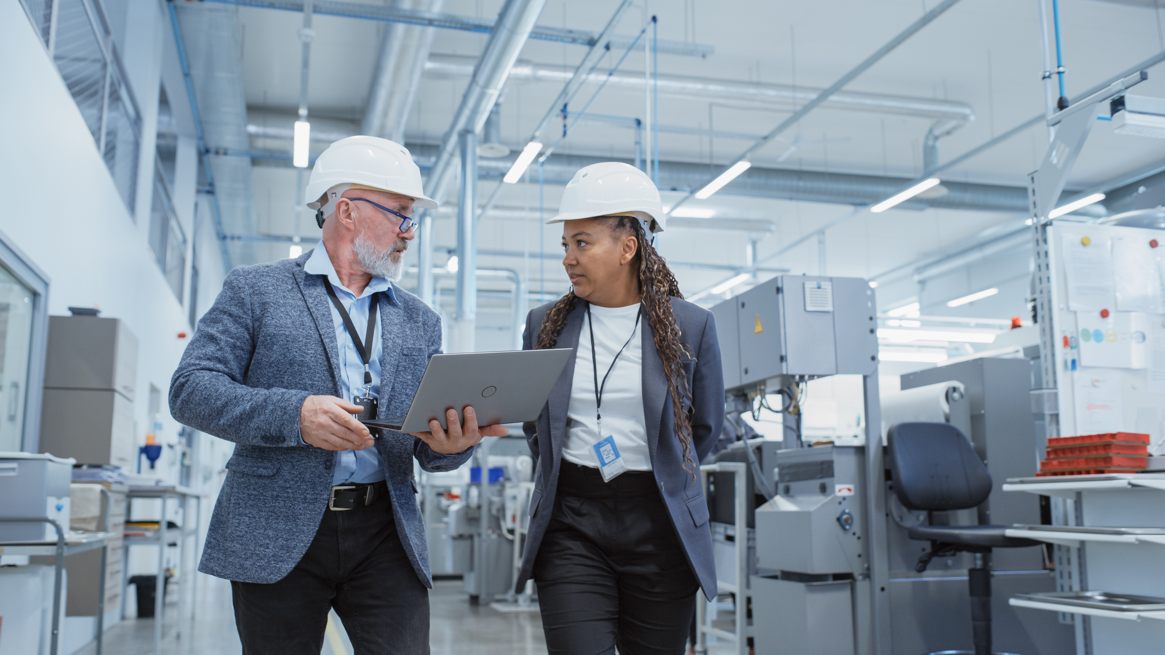 Portrait of a Two Diverse Heavy Industry Manufacturing Engineers in Hard Hats Walking with Laptop Computer and Talking in a Factory. Footage of Two Manufacturing Employees at Work.